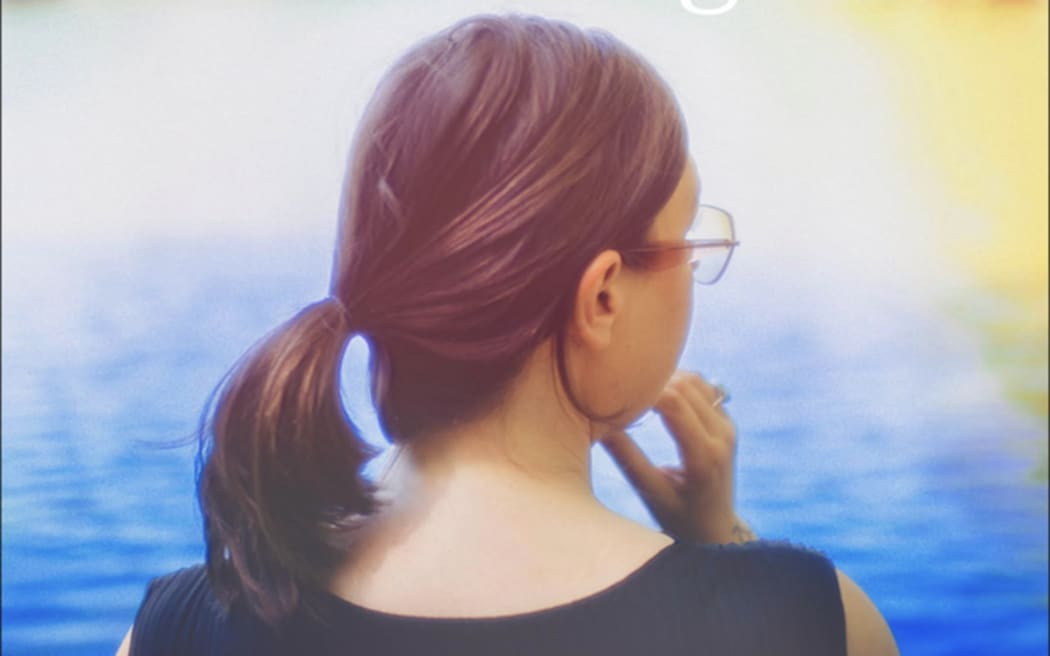 Nadia Reid, a woman with a brown ponytail and glasses, looks toward some blue water. NZ folk singer Nadia Reid's 2025 album Enter Now Brightness