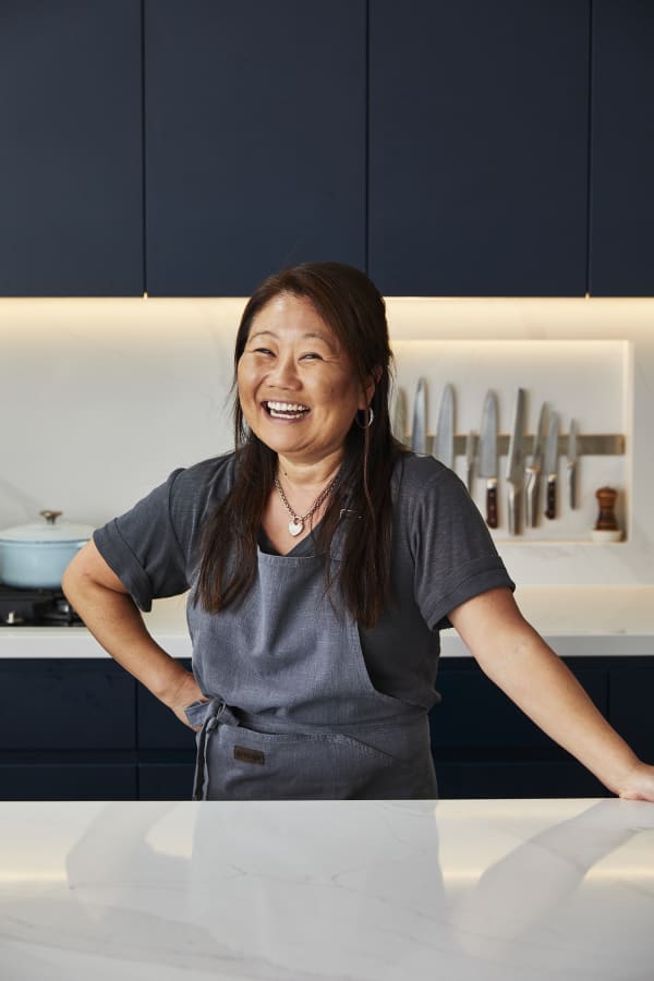 Nagi stands in her kitchen, smiling, wearing a grey apron. Behind her on the kitchen bench is a crockpot, and an assortment of chef's knives.