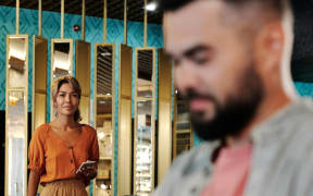 A woman in an orange top carrying a phone approaches a bearded man.