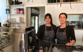 Two women, Keita Powley and Siza Avakh, stand side by side behind the counter in a coffee shop. They are wearing black tops and aprons.