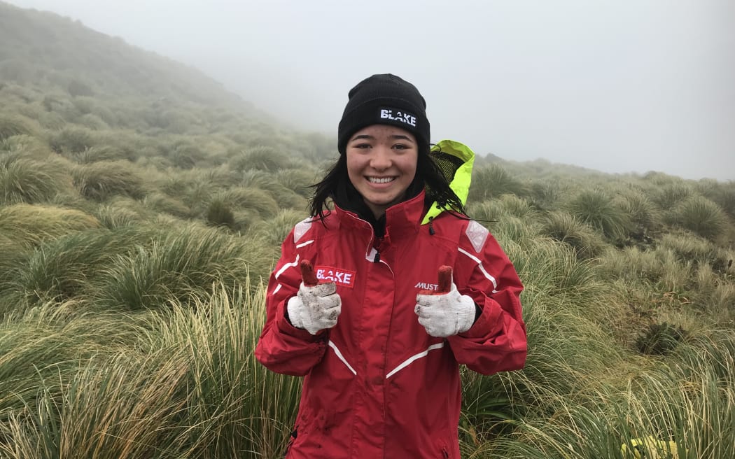 A young lady stands in the tussock in a red Blake jacket, black Blake hat, with white and red gloves up. She is smiling and holding two thumbs up!