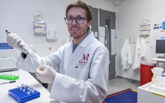A man in a white lab coat and rubber gloves holds a pipette up to an eppendorf tube in a lab.