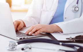 Unknown woman-doctor typing on laptop computer while sitting at the table in sunny clinic.