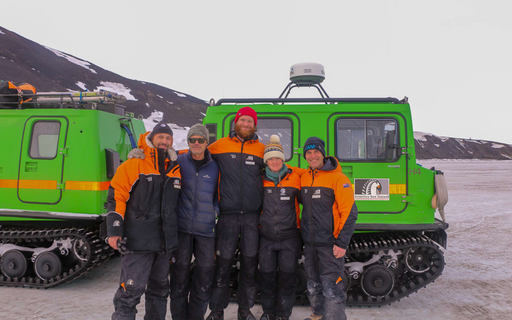 Five people in orange and black thick jackets and beanies stand in front of a bright green vehicle with heavy-duty all-terrain tracks that look like a bulldozer's tracks. A rocky, snow-strewn slope is visible in the background beneath a grey overcast sky.