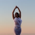 The album cover for 'Sweet Surrender'. Singer/songwriter Aaradhna poses against the background of the setting sun at the beach. Her arms in the air.