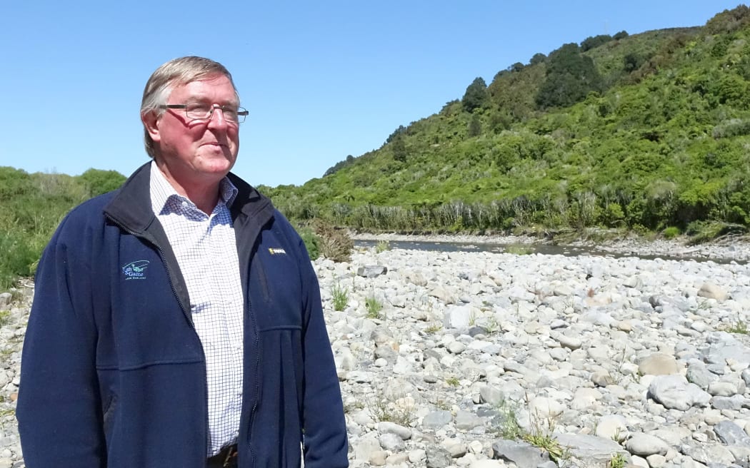 Bryce Johnson  stands on rock bank with bush on other side of the river