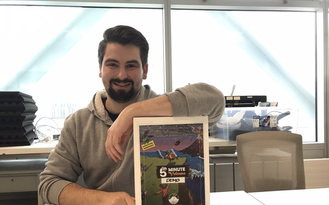 A man with dark hair and a beard sits at a table with his forearm draped over a poster in a frame which says '5 minute volcano demo' and features a cartoon kiwi wearing a hard hat and high vis running away from an erupting volcano.