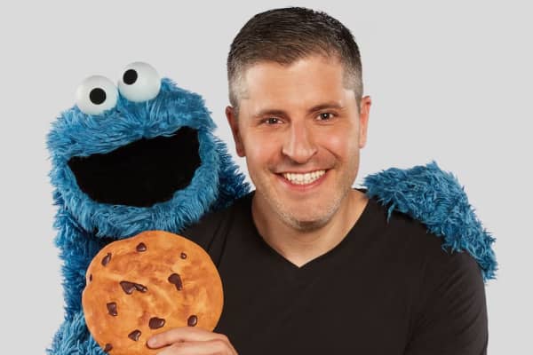 A smiling man holds a cookie and hugs a blue puppet.