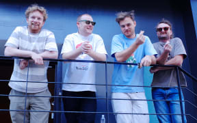 Four men wearing t-shirts stand in a row on a balcony.