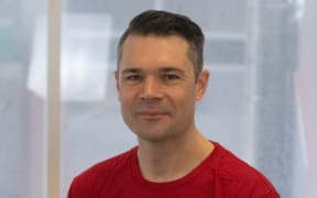 This is a head shot of Tristan. He is sat in front of a window and wearing a red t-shirt that reads "optimise your ride".