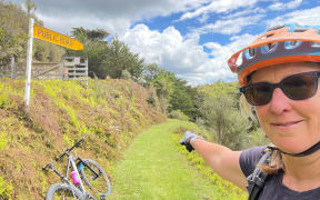 Lynley is wearing sunglasses and a bike helmet. She is standing on the right hand side of the photo and pointing down a grass cycle trail. You can see her bike lying against a grass bank in the background.