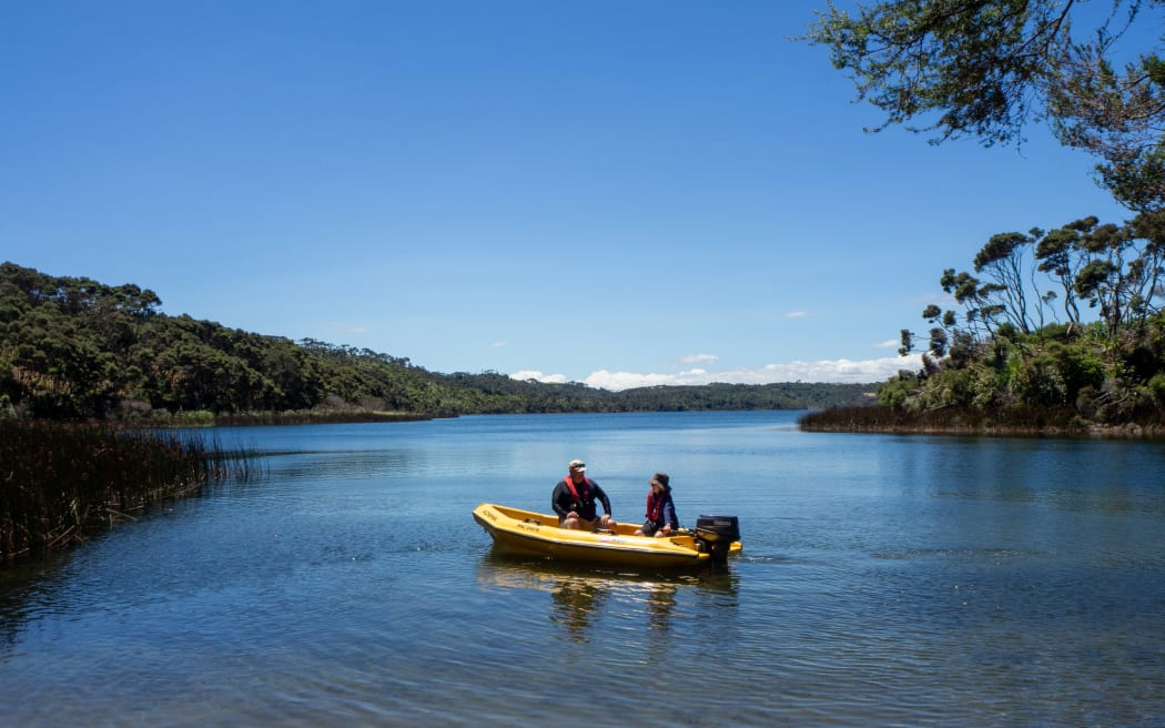 Two people in a small yellow motorised dinghy in the shallows of a wide blue lake fringed by bush beneath a blue sky.