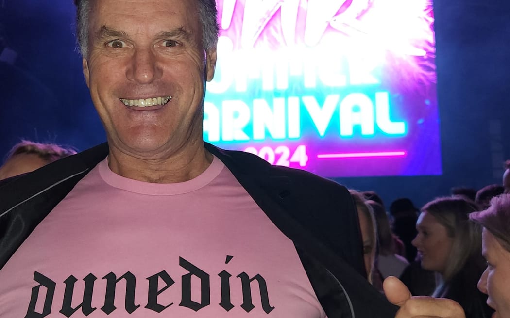 Jules Radich smiles broadly at the camera. He holds his jacket open to show a pink tshirt with the gothic logo "DUNEDIN". He is standing in the mosh of the Pink concert. There are large bright screens behind him, and hundreds of people.