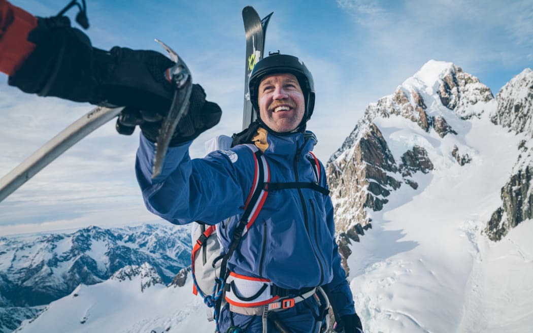Sam Smothy stands on the side of a mountain. He has an ice axe in one hand and his ski's strapped to his back.