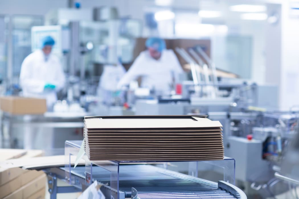 Workers on production line in apharmaceutical plant, cardboard boxes stacked in foreground.