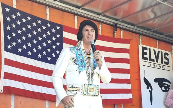 An Elvis impersonator stands on a stage on the back of a truck with an American flag stretched out on the wall behind him.