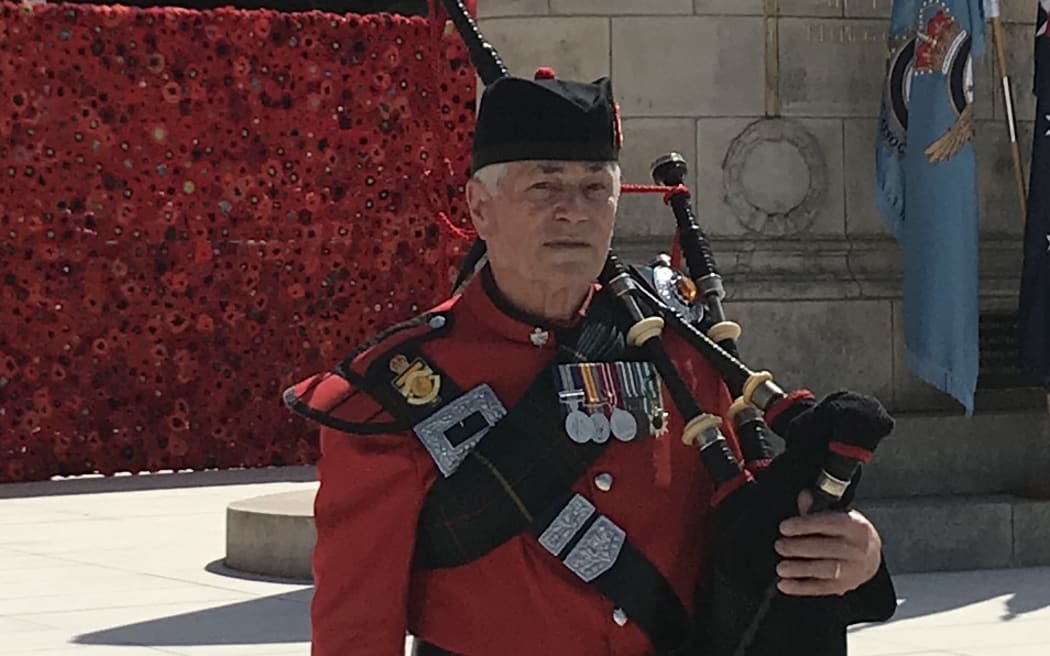 Les Kenfield is standing in front of a war memorial. He is in a kilt and red military jacket. He is holding his bagpipes.