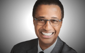 Harold smiles at the camera. He wears a grey suit jacket with a black and white polka dot tie.