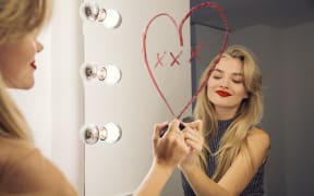 A woman looking in a bathroom mirror drawing a heart on the mirror with lipstick. (Photo by Manuela / Cultura Creative / Cultura Creative via AFP)