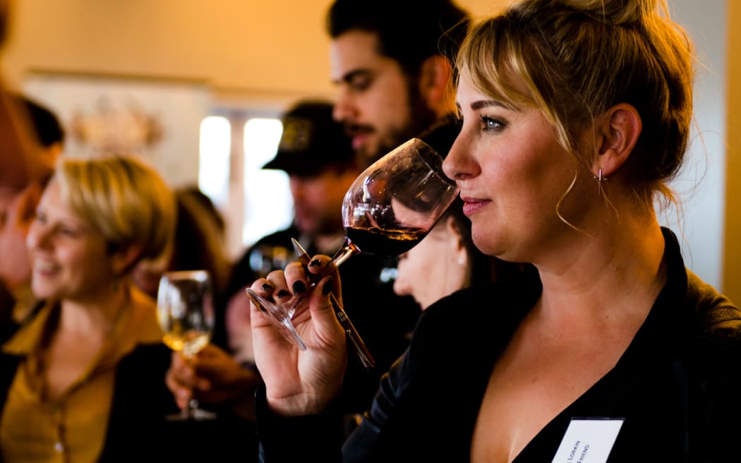 Yvonne lifts a glass of red wine to her nose. She looks off into the distance. She is standing at a wine tasting event indoors.