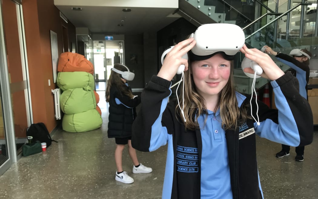 A student lifts up a virtual reality headset so you can see her face. She is holding two controllers. Behind her are two other students with headsets on.