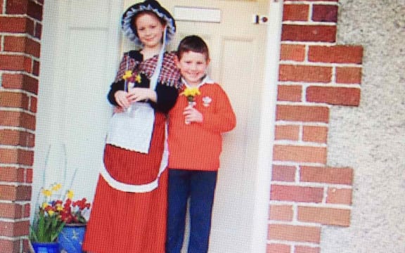 Two children stand on the steps outside the front door to their house. They are wearing red and holding daffodils.