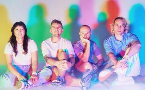 The Beths - a woman with long dark hair and four men - sit in a row with colourful shapes on the wall behind them.