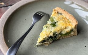 A slice of quiche on a plate. Green beans and spinach is visible among the creamy egg mixture.