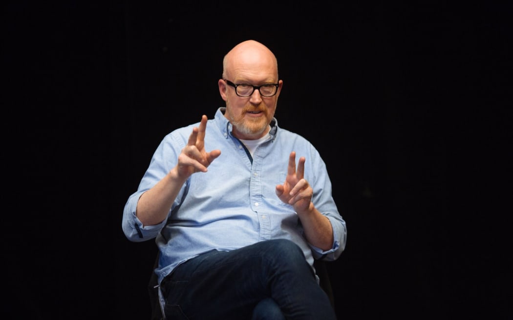 Joe stands on a stage gesturing and speaking against a black background.