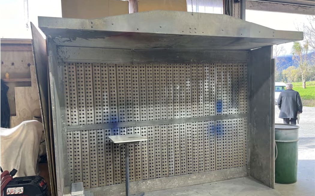 A large silver stand, pictured in the Gloriavale paint shop, where some aerosol paint spraying is done. In the back ground, a roller door to outside and a large drum can be seen.