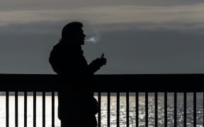 A black-and-white image of a silhouetted person vaping in front of a fence adjacent to a body of water.