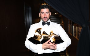 Bad Bunny, wearing a white shirt and black bowtie, bundles three golden trophies in his arms.