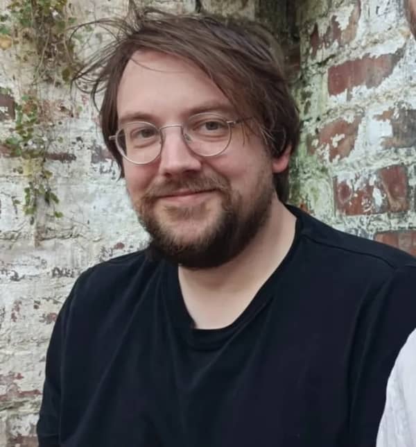 Tom Whyman is a bearded man wearing round wire-rimmed glasses and a navy blue t-shirt.