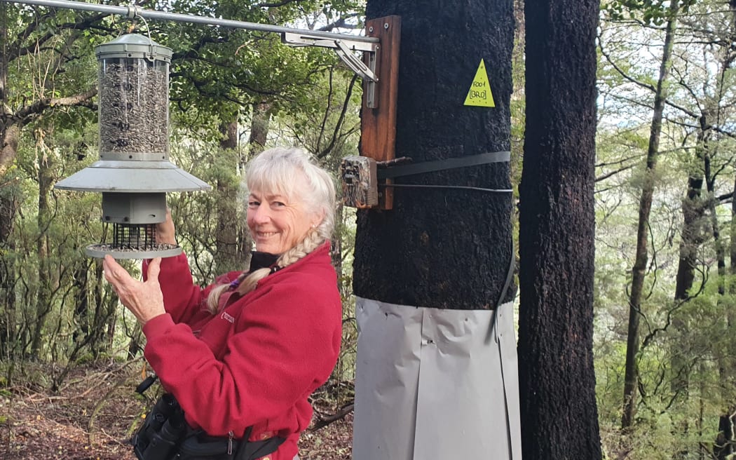Neroli Amyes is looking towards the camera, smiling, wearing a red jacket. She is holding a large bird feeder which is hanging off a metal cable attached to large pole.