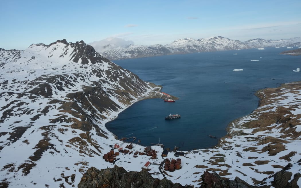 Looking down from a high mountain onto a bay surrounded by snowy, rocky hills. The bay is dark blue and looks cold. There is a ship sitting in the middle of the bay and icebergs visible further out.
