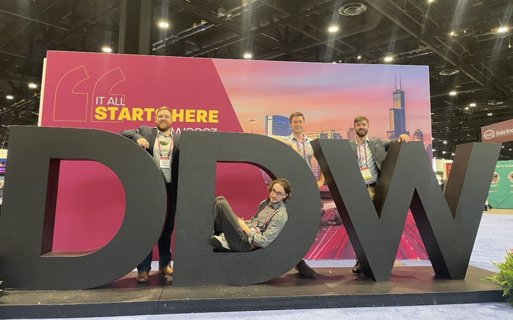 Peter and three other men pose around a sculpture of three large letters, "DDW", meaning Digestive Disease Week, inside a large conference hall.