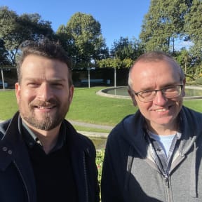 Rob and Chris smile at the camera. They're standing in a park.