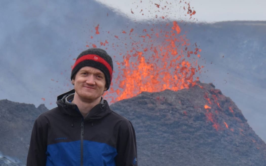 Dr Oliver Lamb, dressed in winter outdoor clothing, stands in front of a volcano erupting lava in Iceland