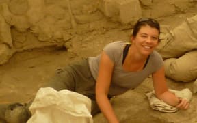 Dr Eline Schotsmans is laying on the ground digging a hole. The contents of the hole are out of shot but the surroundings imply it will be bones. She is looking towards the camera and smiling.