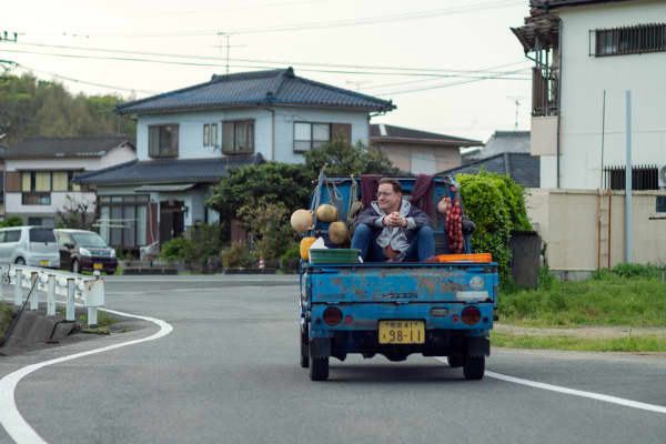 Brendan Fraser as Phillip Vanderploeg on the back of a blue truck carrying veggies and buckets in the film Rental Family.