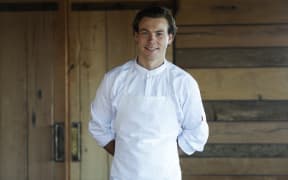 Jack Cashmore wears chef whites and stands infront of a "rustic" looking wooden door at Blue Duck Station.