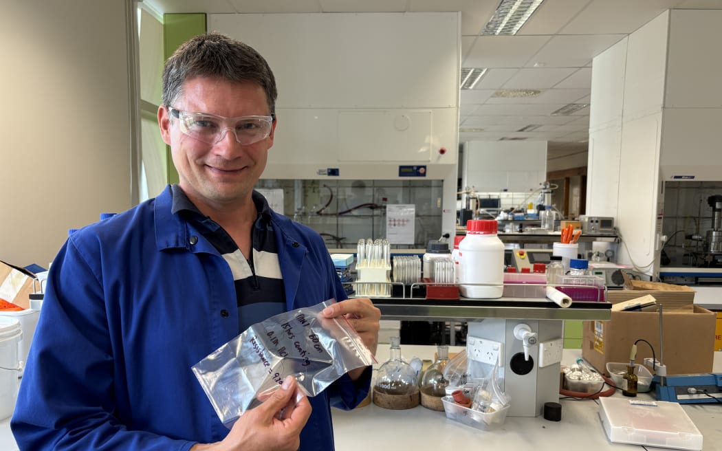 Robert is standing in a chemistry lab beside a bench, a fume hood is visible in the background and in the foreground there are chemicals on the benchtop. He's wearing a blue lab coat and clear safety goggles. He's smiling at the camera, holding a small clear plastic bag with writing on it.