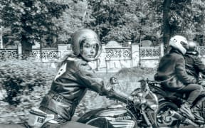 A woman on a motorbike looks at the photographer with her hair flying out behind her.