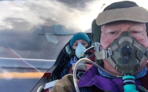 Two people, both wearing oxygen masks, in the cockpit of a glider.