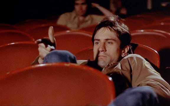 Movie still from Martin Scorsese's film Taxi Driver showing Robert De Niro as Travis Bickle slouching in a cinema with his fingers pointing like a gun barrel.