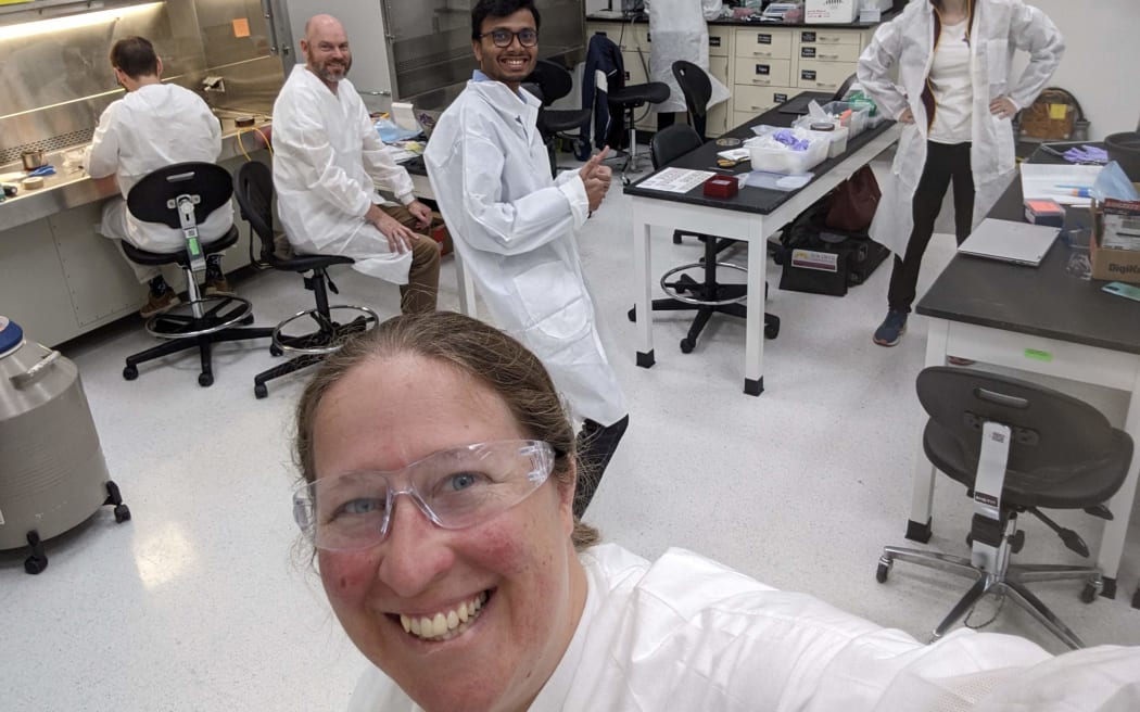 A smiling person wearing a white lab coat and safety glasses takes a selfie in a laboratory setting with several others, also wearing white lab coats. Some are seated at workstations, while others are standing or walking around. The lab is equipped with various scientific instruments and equipment, including a fume hood, cabinets, and tables with supplies.