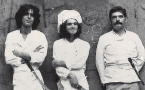 Three men wearing white aprons and carrying knifes are lined up against a wall in a black and white photo.