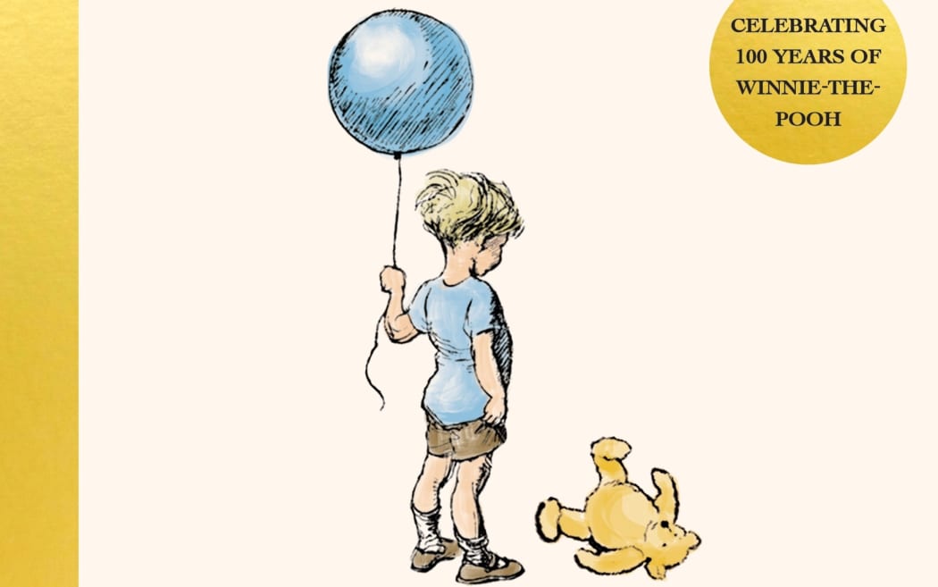 A small boy holds onto a blue balloon and looks down at his yellow toy bear at his feet