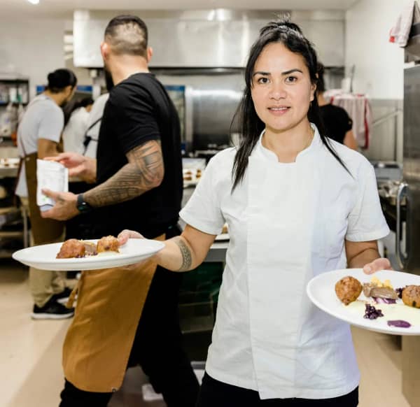 Tongan-born chef Ana Mailei Savio, of Onehunga-based online meal service Gia's Grab&Go, holds two plates of food in a busy kitchen.