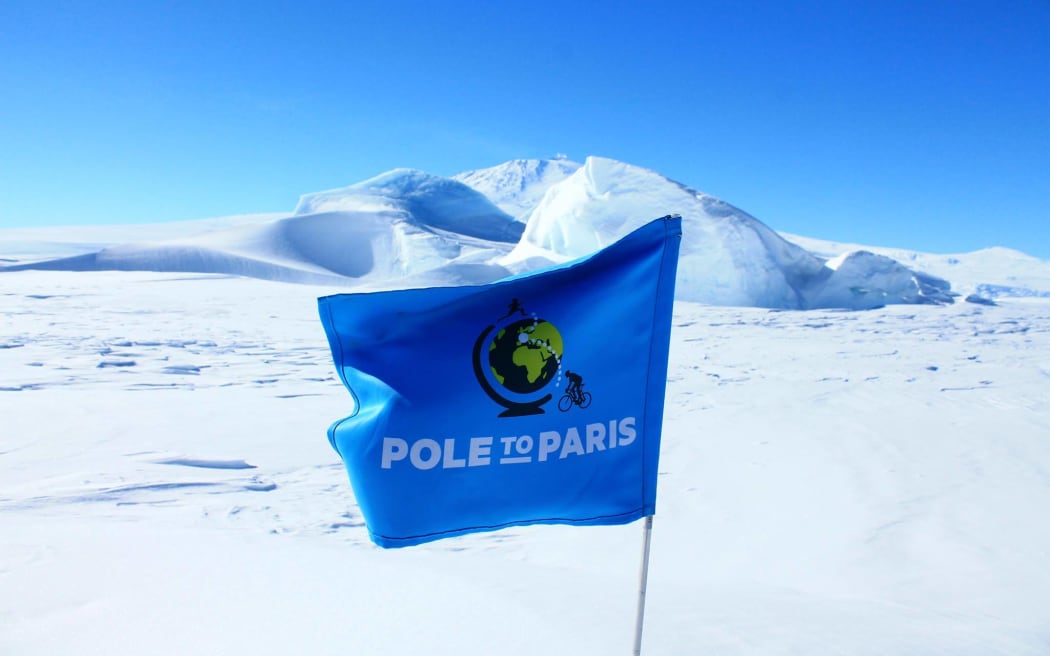A blue flag planted in the snow reads "Pole to Paris". In the background, a hill covered in ice and snow beneath a bluebird sky.